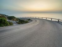 an empty road curves into the distance of some mountains with ocean in background and sun setting