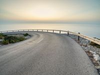 an empty road curves into the distance of some mountains with ocean in background and sun setting