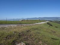 an empty road with mountains in the distance while two horses graze on grass behind it