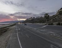 the motorcycle is going down the empty road near the beach at dusk, along with palm trees and mountains