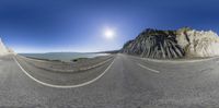 a fish eye shot of an empty road near some mountains and water with rocks in the background