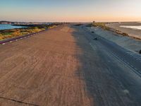 an empty road next to the ocean during sunset and in the foreground there are cars driving along the coast