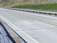 an empty road near some green hills and trees on the side of a bridge with no traffic