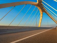 an empty road underneath a bridge with yellow arches and cables and some blue sky in the background