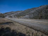 an empty road that is near a rocky hill with trees growing up behind it on a clear blue sky day