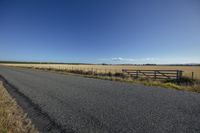 a empty road runs through an open grassy area to the horizon, as clouds above a fence