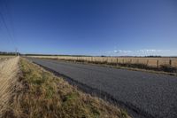 an empty road with no traffic on it, next to a rural field with dry grass and electric wires