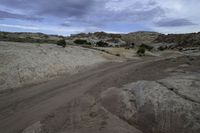 an empty road running through some dirt field near a rock formation and tree line in the distance