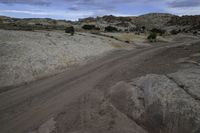 an empty road running through some dirt field near a rock formation and tree line in the distance