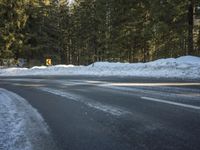 an empty street covered in snow and snow next to a forest with lots of trees