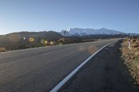 a long stretch of empty road with snow covered mountains behind it in the distance on a sunny day