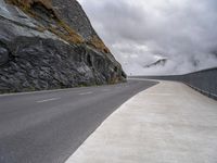 the empty road runs between rock walls and a cliff wall below it is covered in clouds