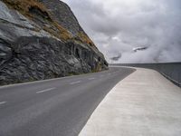 the empty road runs between rock walls and a cliff wall below it is covered in clouds