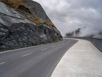 the empty road runs between rock walls and a cliff wall below it is covered in clouds