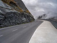 the empty road runs between rock walls and a cliff wall below it is covered in clouds