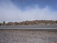 an empty road surrounded by rocks and shrubs on a bright cloudy day with clouds in the blue sky