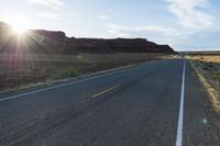 an empty road with sun flareing out of it near the mountains at sunset,