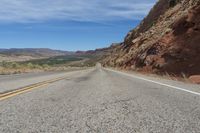 an empty road in the middle of the desert with mountains behind it and a red rock cliff in the distance