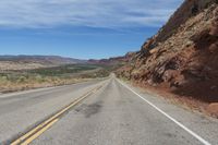 an empty road in the middle of the desert with mountains behind it and a red rock cliff in the distance