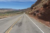 an empty road in the middle of the desert with mountains behind it and a red rock cliff in the distance