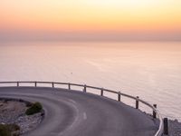 an empty roadway on a cliff overlooking the ocean at sunset with a bird flying over the edge