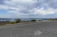 an empty roadway near the coast near some trees and water in the distance is seen through clouds