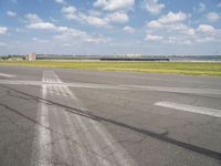 an empty runway next to grassy field with clouds in the sky over it and some buildings in the distance