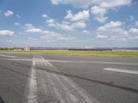 an empty runway next to grassy field with clouds in the sky over it and some buildings in the distance