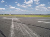 an empty runway next to grassy field with clouds in the sky over it and some buildings in the distance