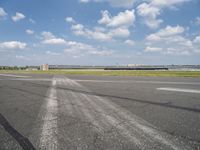 an empty runway next to grassy field with clouds in the sky over it and some buildings in the distance