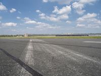 an empty runway next to grassy field with clouds in the sky over it and some buildings in the distance