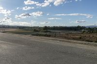 an empty street with a lot of trees in the distance and clouds above it and some dirt ground in the foreground