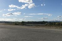 an empty street with a lot of trees in the distance and clouds above it and some dirt ground in the foreground