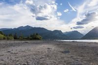 an empty sandy beach in a mountainous area under a blue sky with clouds and a mountain range