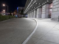 an empty sidewalk in a large building area at night, with a bench and a bicycle rack attached to the side