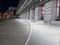 an empty sidewalk in a large building area at night, with a bench and a bicycle rack attached to the side