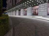 an empty sidewalk in a large building area at night, with a bench and a bicycle rack attached to the side