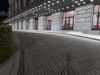 an empty sidewalk in a large building area at night, with a bench and a bicycle rack attached to the side