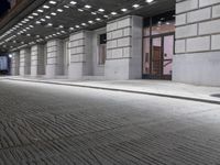 an empty sidewalk in a large building area at night, with a bench and a bicycle rack attached to the side