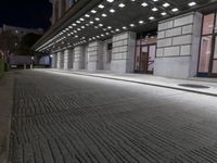 an empty sidewalk in a large building area at night, with a bench and a bicycle rack attached to the side
