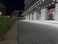 an empty sidewalk in a large building area at night, with a bench and a bicycle rack attached to the side