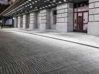 an empty sidewalk in a large building area at night, with a bench and a bicycle rack attached to the side