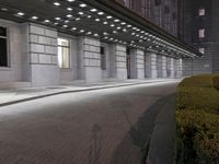 an empty sidewalk in a large building area at night, with a bench and a bicycle rack attached to the side