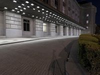 an empty sidewalk in a large building area at night, with a bench and a bicycle rack attached to the side