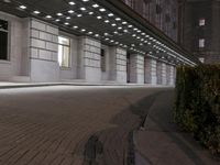 an empty sidewalk in a large building area at night, with a bench and a bicycle rack attached to the side