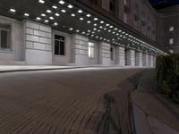 an empty sidewalk in a large building area at night, with a bench and a bicycle rack attached to the side