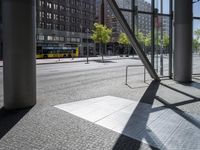 an empty sidewalk in front of tall buildings and a glass enclosure in the background is a brick walkway and a bench on the sidewalk