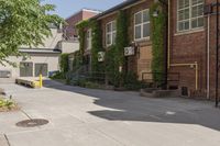 an empty sidewalk between two brick buildings and a paved sidewalk in the foreground, with trees and plants