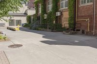 an empty sidewalk between two brick buildings and a paved sidewalk in the foreground, with trees and plants