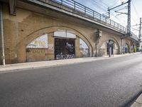 the empty street is next to a brick building and a bridge that has graffiti on it